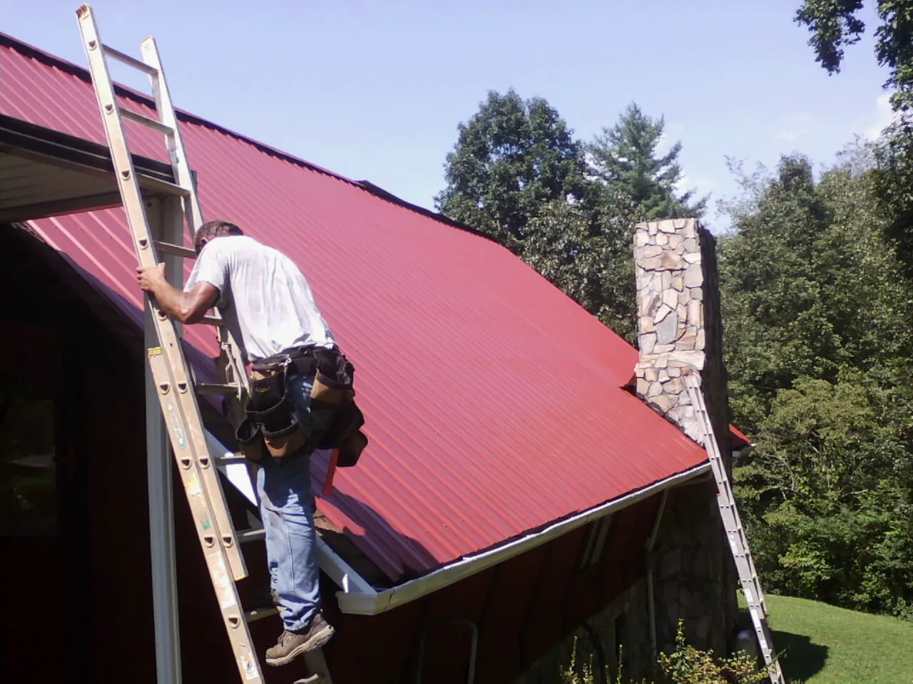 Roofer on a ladder beside a red metal roof and stone chimney — Soffit & Fascia Repair by Roofing Brothers in Coffeen